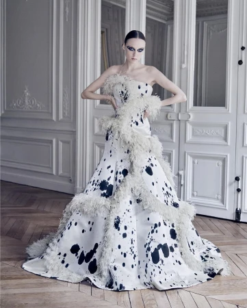 A fashion model poses indoors wearing a white, strapless gown with black abstract spots and white feather trim in front of a paneled wall.
