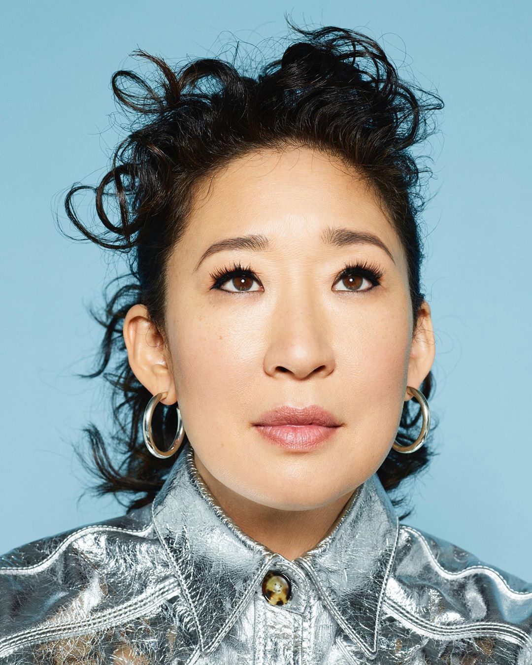 Close-up portrait of Sandra Oh.  She wears a metallic silver collared shirt, hoop earrings and has short dark curly hair, against a light blue backdrop.