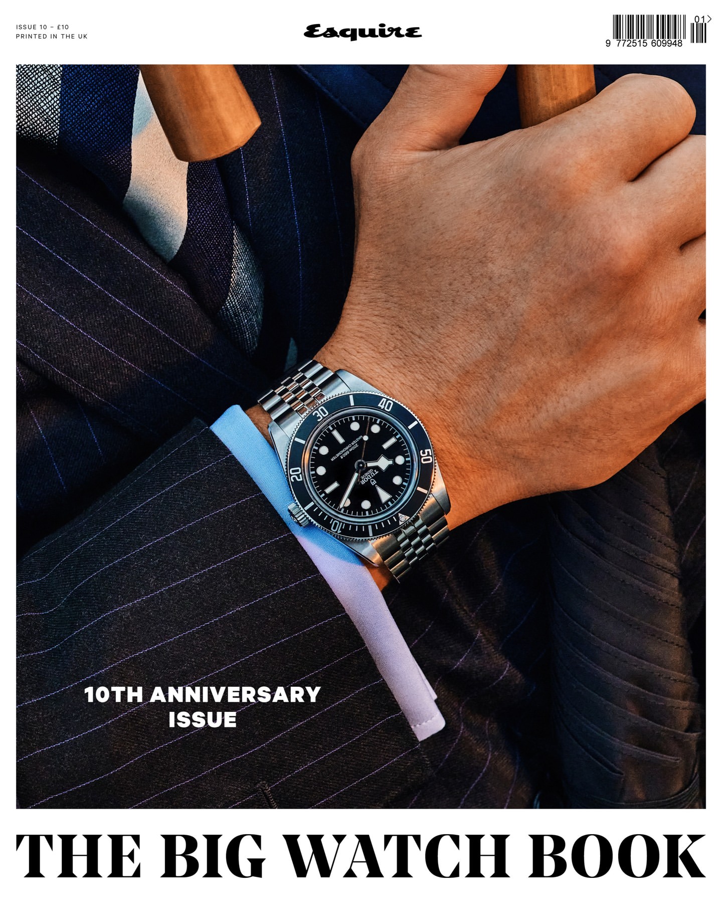 Close-up of a person wearing a steel wristwatch with a black dial and jubilee bracelet, dark pinstripe suit, and light blue shirt.