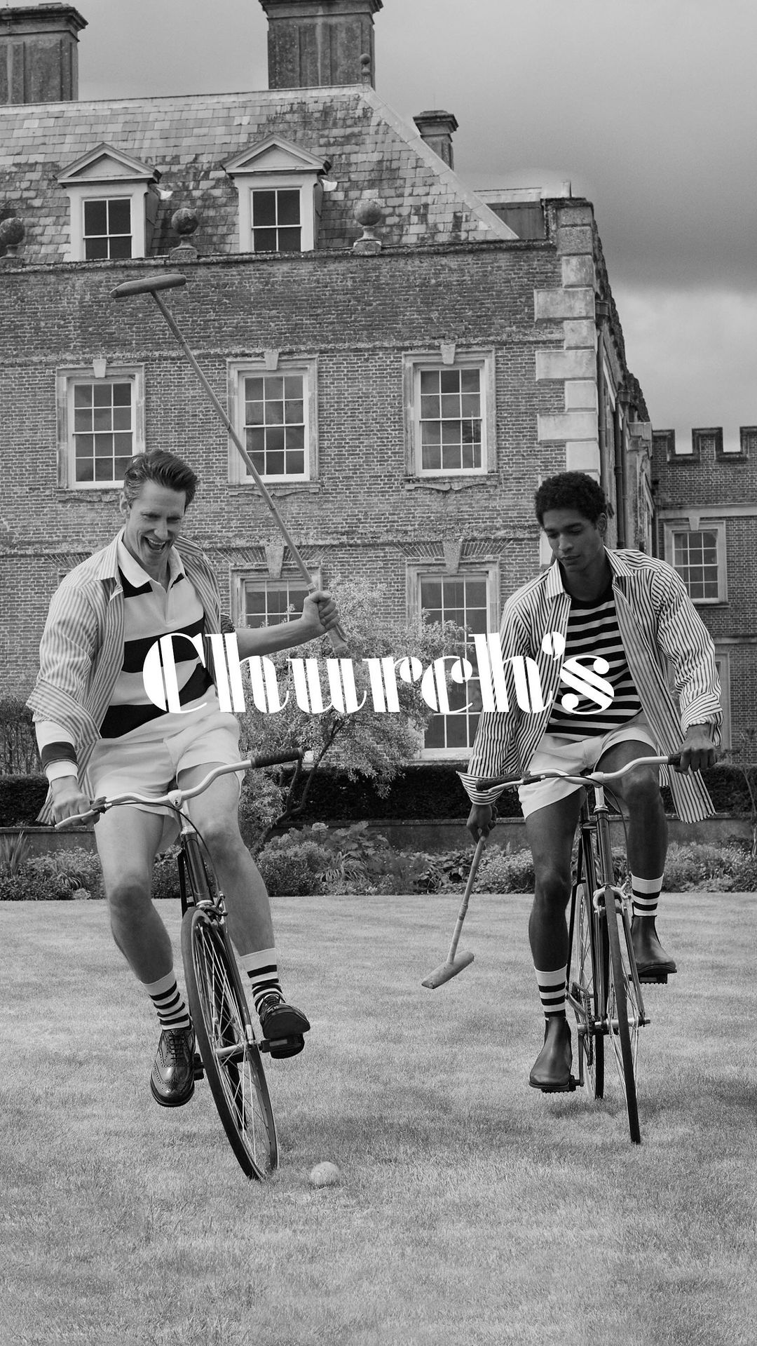 Two adult males ride polo bikes on grass in front of a brick building during the daytime, photographed in black and white.