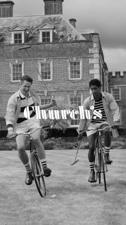 Two adult males ride polo bikes on grass in front of a brick building during the daytime, photographed in black and white.