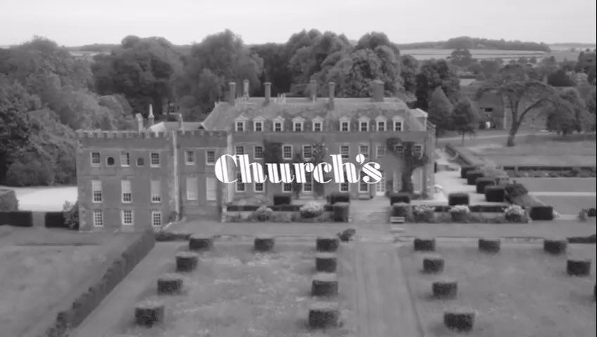 Monochromatic, eye-level shot of a grand manor house with trees in the background, complemented by perfectly manicured, geometrically arranged hedges.