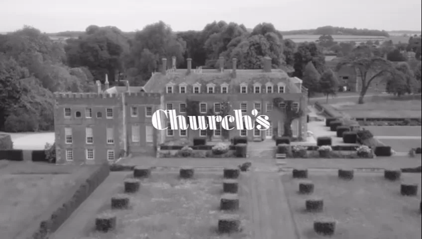 Monochromatic, eye-level shot of a grand manor house with trees in the background, complemented by perfectly manicured, geometrically arranged hedges.