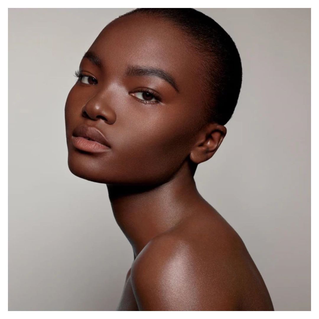 Close-up portrait of a young Black female model with a smooth skin tone, posing in front of a neutral light grey background.