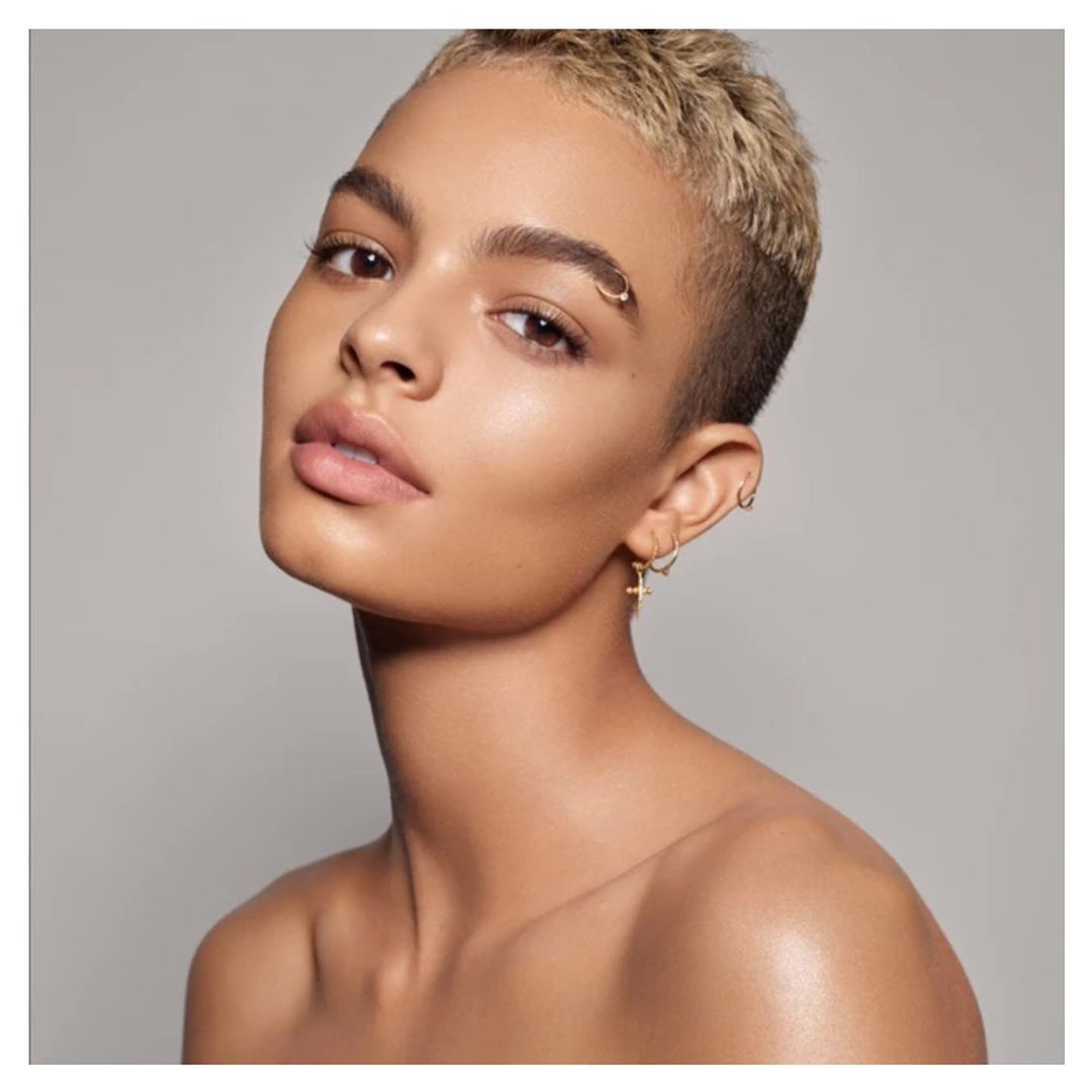 Close-up studio portrait of a young, light-skinned adult female with short blonde hair, neutral expression, and gold jewelry against grey background.