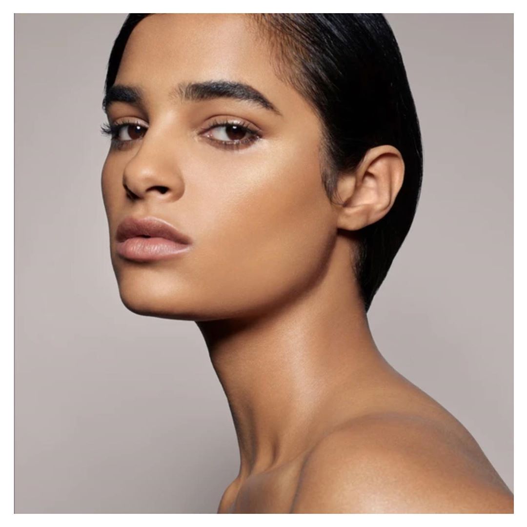 Close-up portrait of a tan-skinned woman with slicked back dark hair, subtly highlighting her face with neutral background, beauty shot.