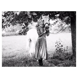 Black and white portrait of a woman in a patterned dress standing next to a white horse under a tree.
