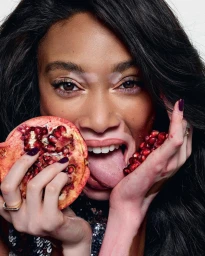 Close-up portrait of Winnie Harlow holding halved pomegranates near her face with dark manicured nails against a white background.