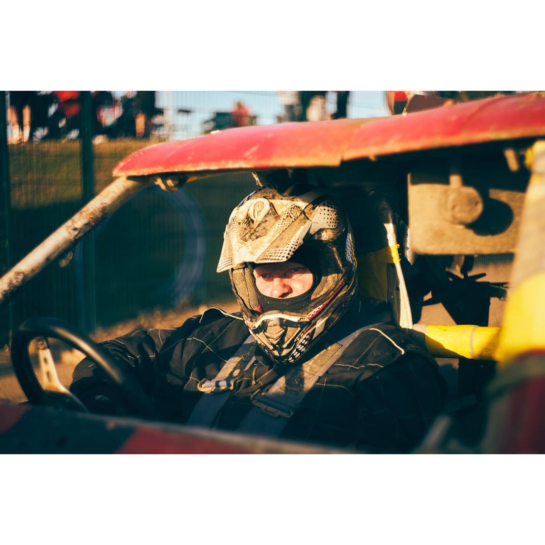 Adult racer wearing helmet drives red, yellow buggy on a dirt track with grassy hills and blue sky in the background.