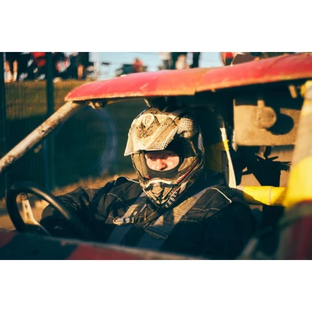 Adult racer wearing helmet drives red, yellow buggy on a dirt track with grassy hills and blue sky in the background.