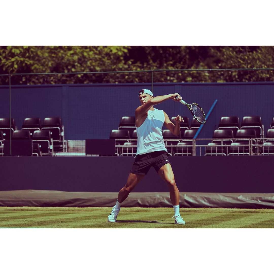 A male tennis player in a white tank top and black shorts is swinging a tennis racket on a grass court with a blue background.