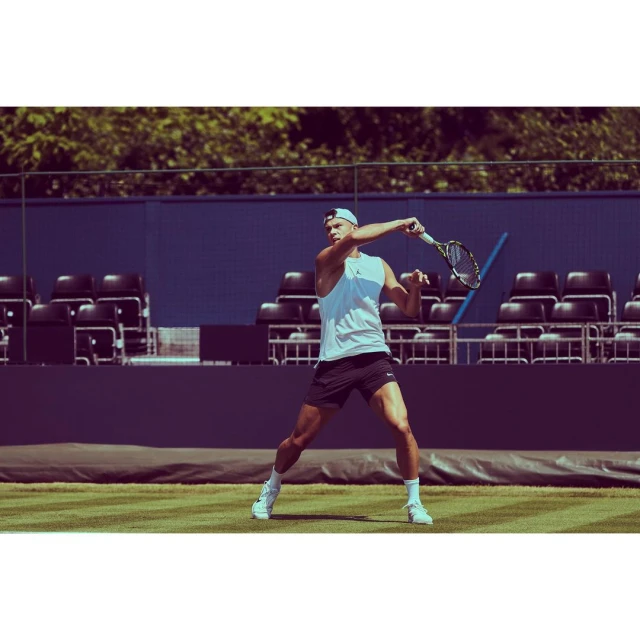 A male tennis player in a white tank top and black shorts is swinging a tennis racket on a grass court with a blue background.