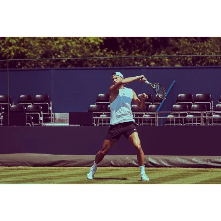 A male tennis player in a white tank top and black shorts is swinging a tennis racket on a grass court with a blue background.
