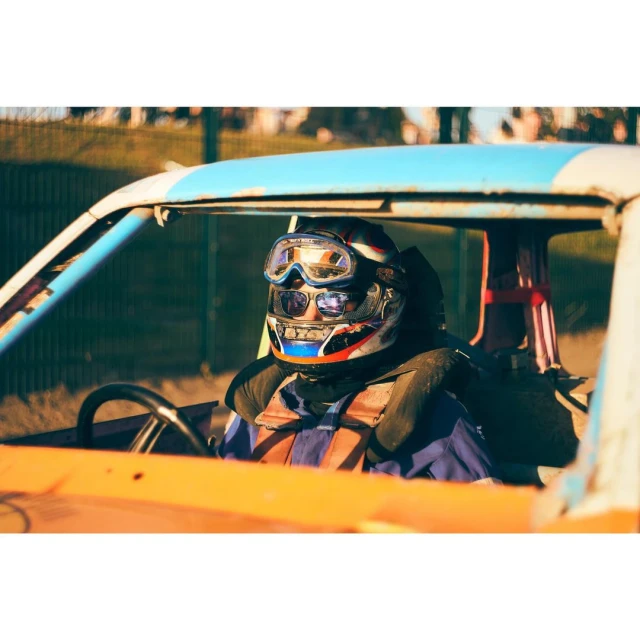 Demolition derby driver in helmet and safety gear sits in damaged car; blue and white frame with orange interior in outdoor setting.