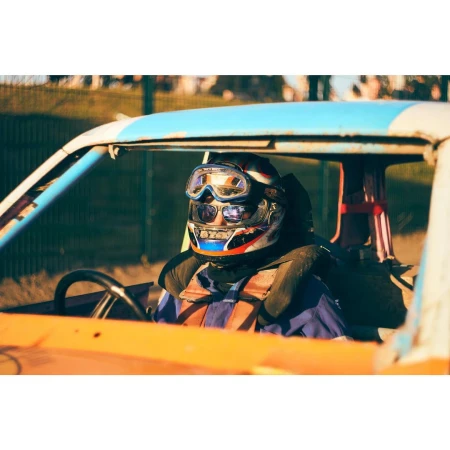 Demolition derby driver in helmet and safety gear sits in damaged car; blue and white frame with orange interior in outdoor setting.