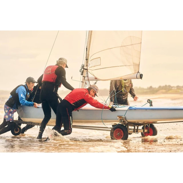 Four people push a sailboat on a wheeled trailer into the water at the beach on an overcast day, with waves lapping the shore.