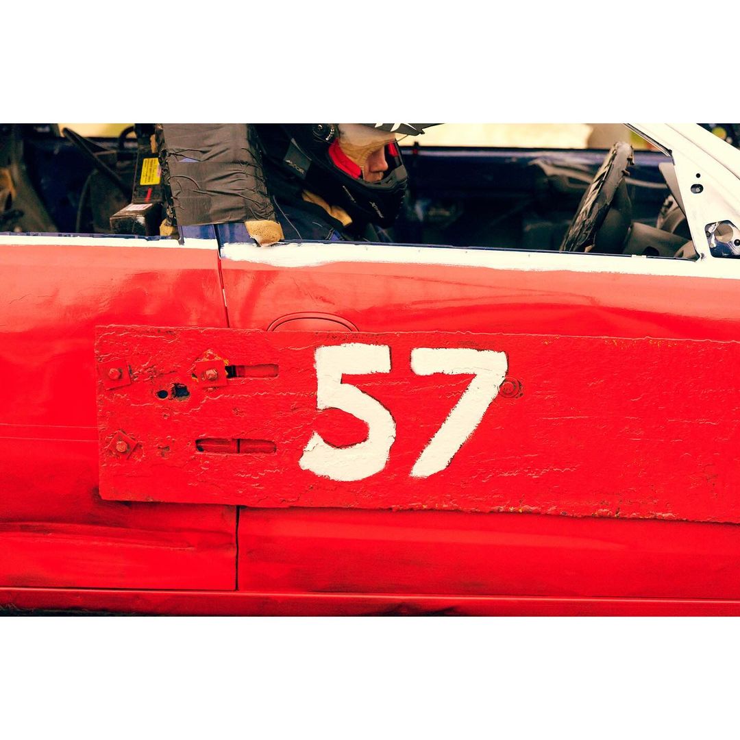 Red race car with the number 57 in white, driver visible in helmet, capturing the excitement of motorsports racing.