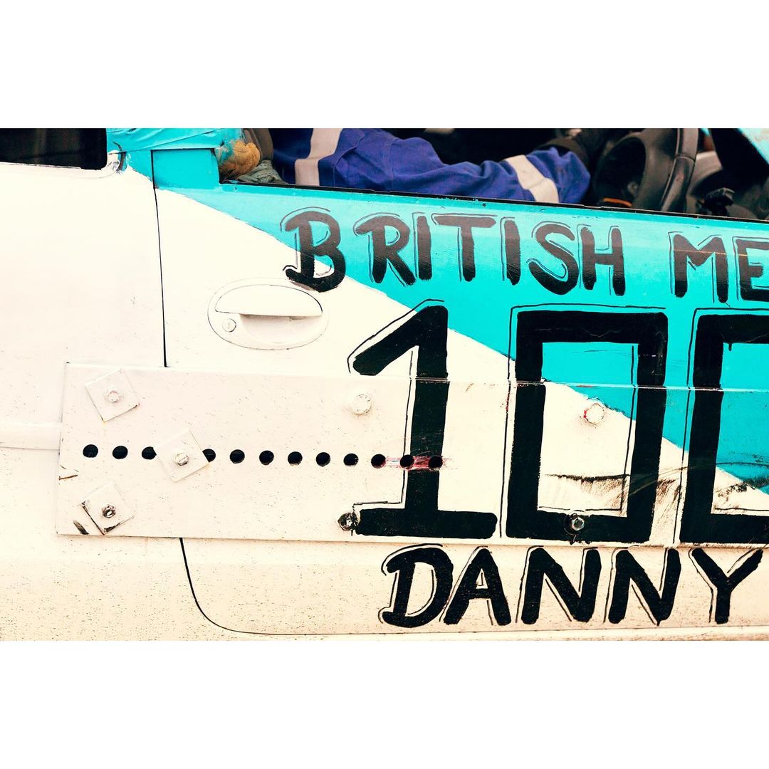 Close up of a white and blue stock car with the number 100 and the name Danny painted on the side, showing racing details.