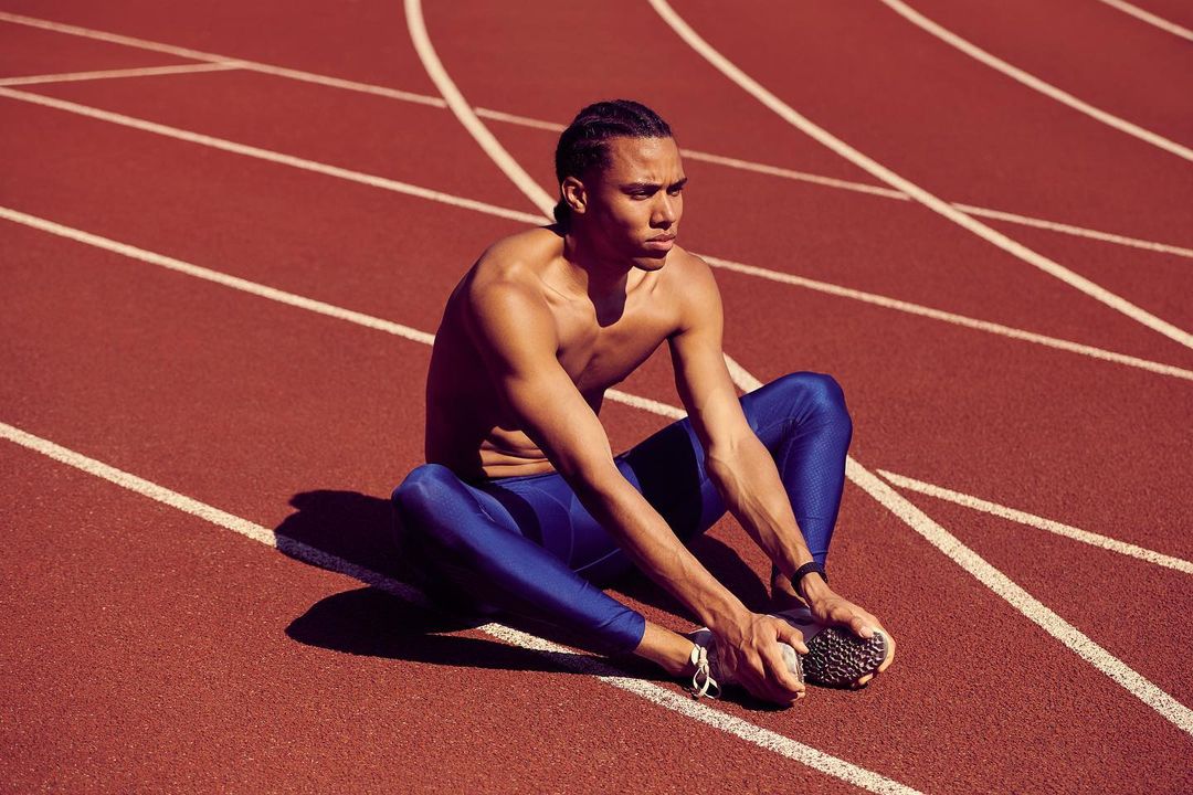 Athlete stretching on a running track, wearing blue leggings, with arms extended to feet and lines of track filling background.