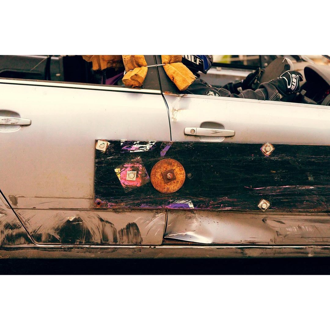 Damaged silver car with a large black panel attached to the side, featuring rust-colored circular elements and taped squares.