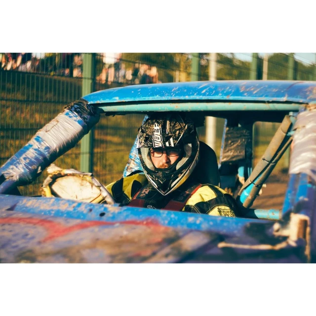 Action shot of a helmeted racer in a blue stock car, safety gear visible, surrounded by an outdoor race track environment.