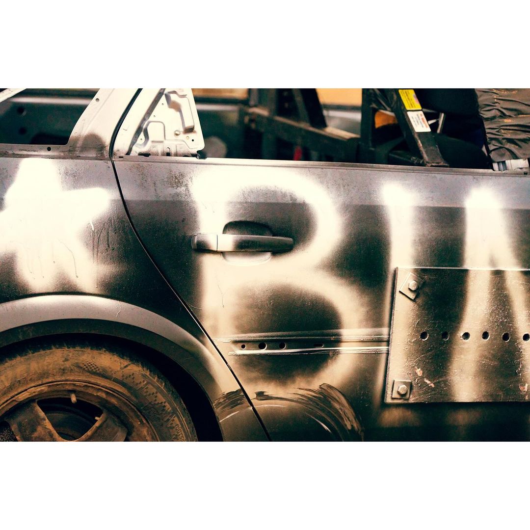 Close-up of a damaged, grey car with white spray paint, showing a flat tire, scratches, and a riveted metal plate.