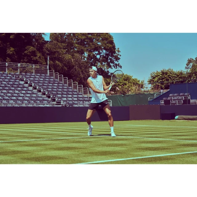 Man in white tank top and black shorts playing tennis on a green court in stadium, sunny day, trees in the background.