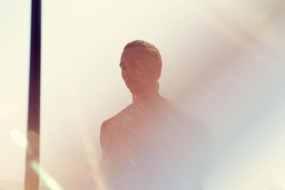 Back view of silhouetted adult under shower, light skin, outdoors. Spraying water, soft diffused light, abstract composition, horizontal format.