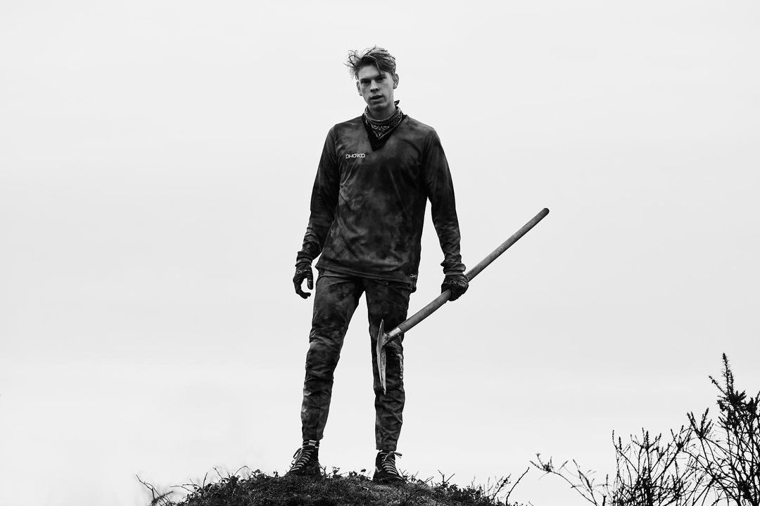 Monochrome portrait of a blonde adult male standing outdoors on a small hill, holding a tool, wearing dark clothing and bandana.