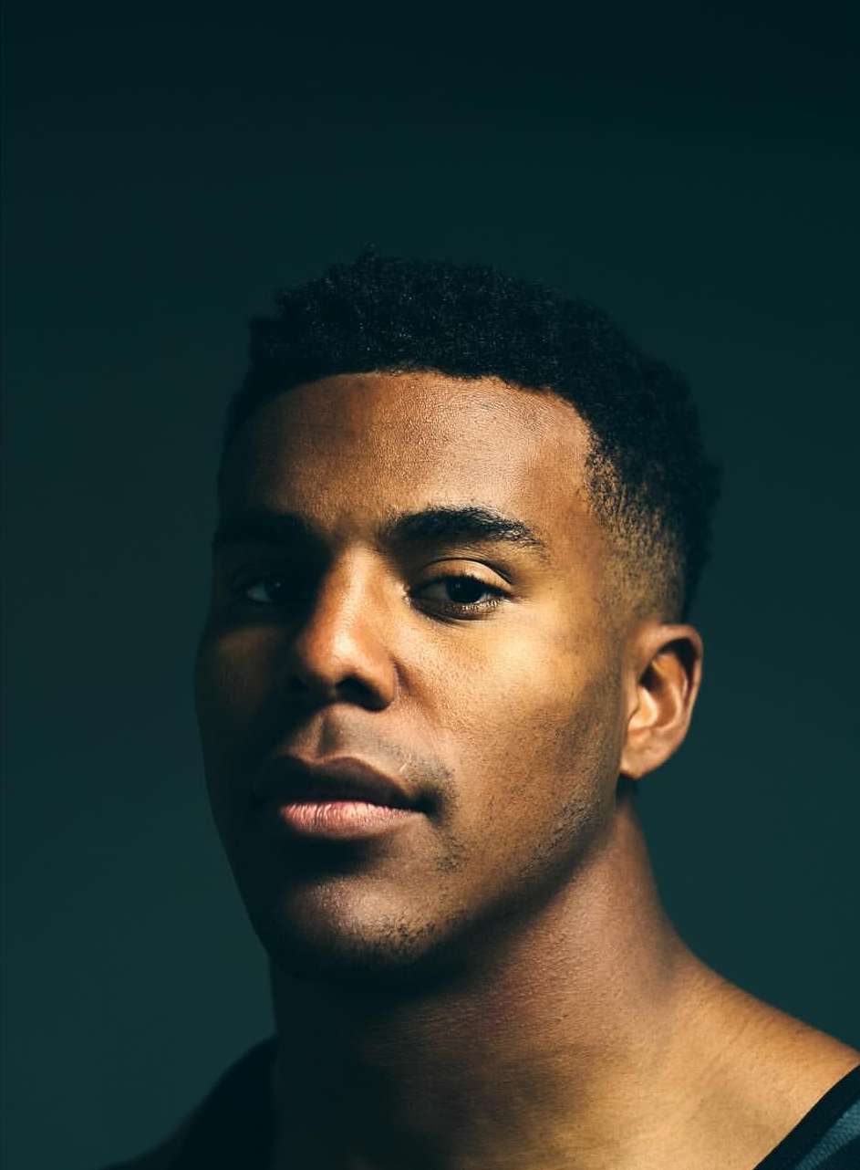 Headshot of an adult African-American man with short, dark hair and a serious expression, photographed in a studio with dark teal background.
