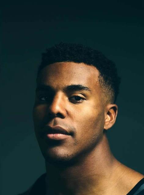 Headshot of an adult African-American man with short, dark hair and a serious expression, photographed in a studio with dark teal background.