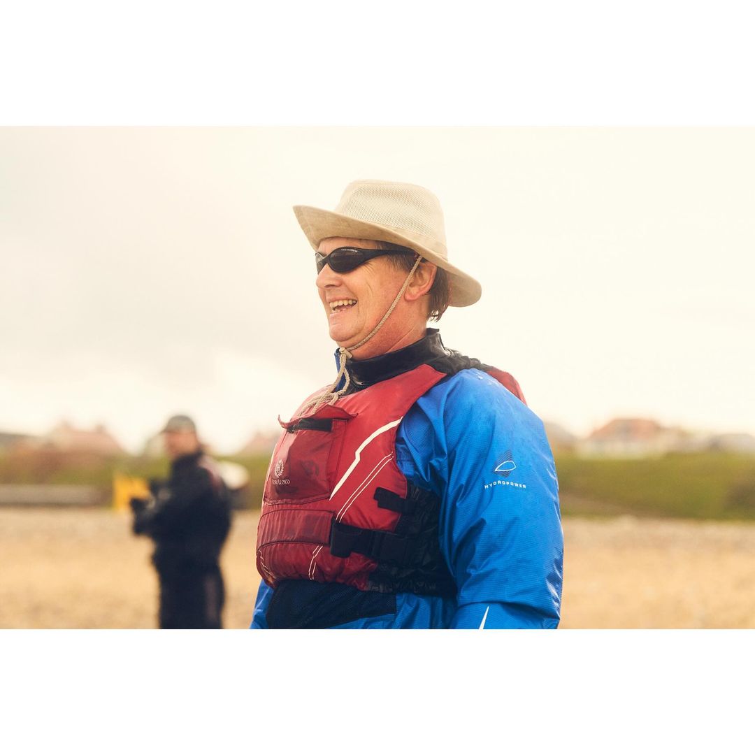 Smiling person on the beach with a life vest and hat, with another person out of focus in the background on a cloudy day.