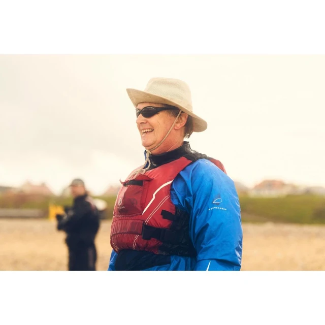 Smiling person on the beach with a life vest and hat, with another person out of focus in the background on a cloudy day.
