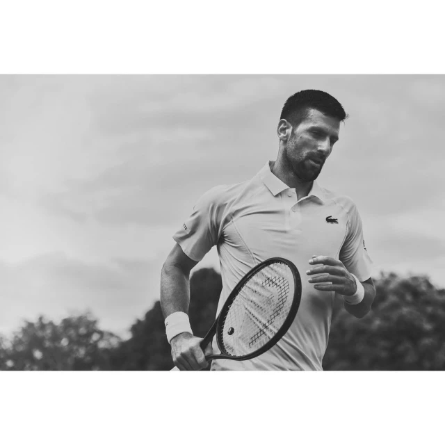 Black and white portrait of a male tennis player holding a racket, outdoors on a court with trees and sky in the background.