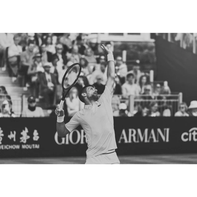 Monochrome photo of a male tennis player in mid-serve on the court with a crowd in the background. He is wearing athletic apparel.