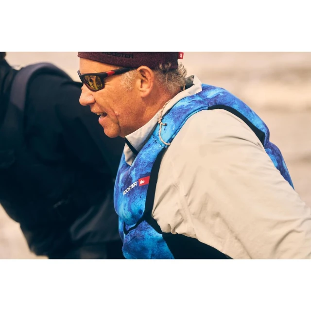 Man wearing a blue and white life jacket with red sunglasses and a burgundy beanie standing outside on what appears to be a beach.