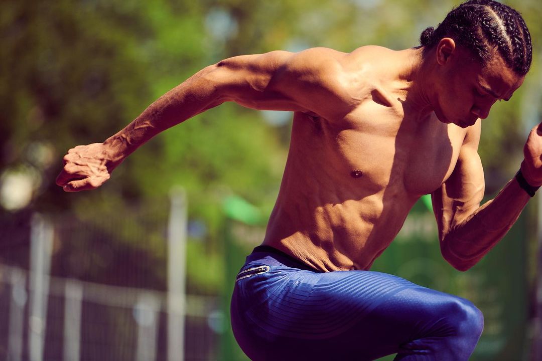 A muscular, shirtless man in blue pants runs outdoors in bright sunlight, showcasing athleticism and fitness against a blurred green backdrop.