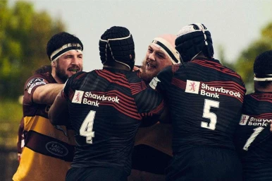 Group of rugby players in striped jerseys with Shawbrook Bank sponsor logos celebrating a try on the pitch