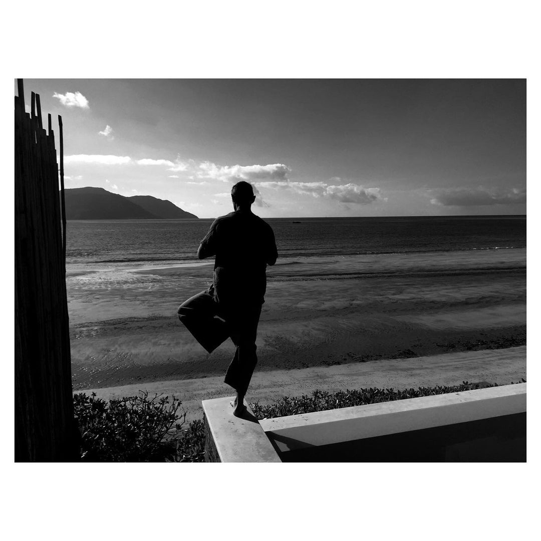 Black and white photograph of a person performing a yoga pose at the beach with ocean and distant mountains in the background under a cloudy sky.