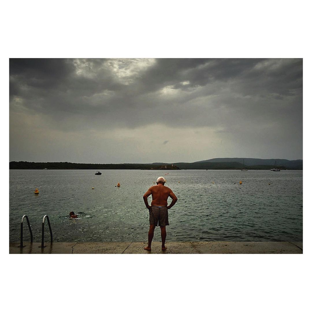 Elderly man facing a lake on a cloudy day, wearing patterned shorts and a white cap, with another person swimming in the distance.