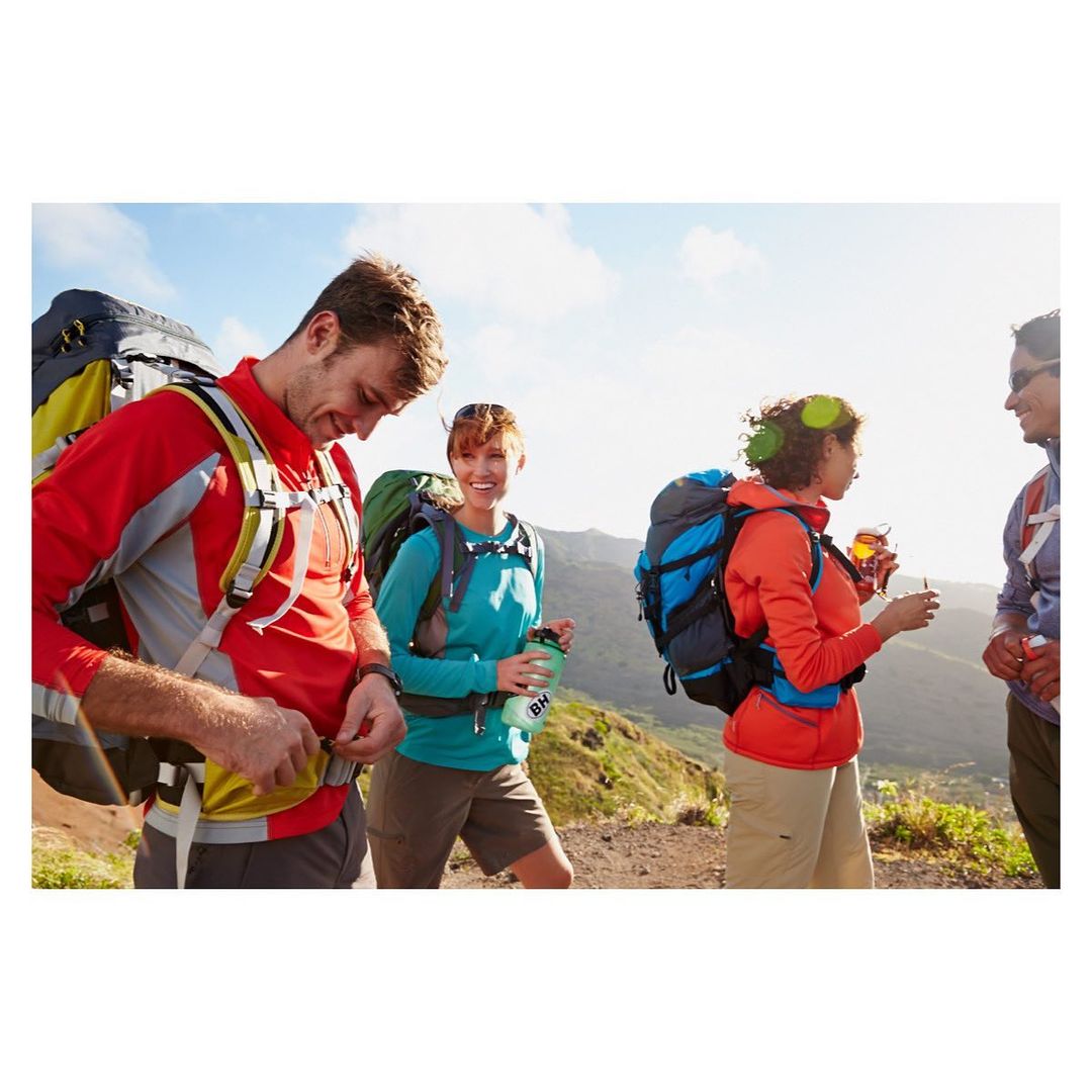 Group of four hikers wearing backpacks on a mountain trail, under a bright sky, chatting and holding water bottles.