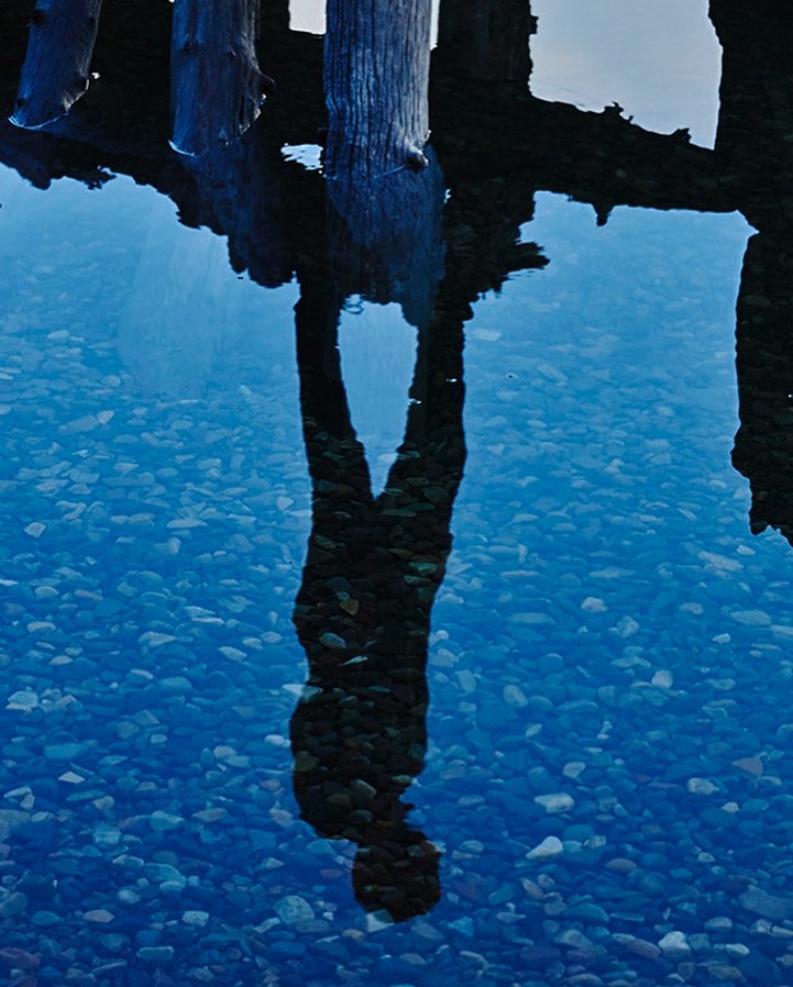 Reflection in clear blue water showing silhouette of person. Rocks visible underwater and wooden pier structure above the surface.