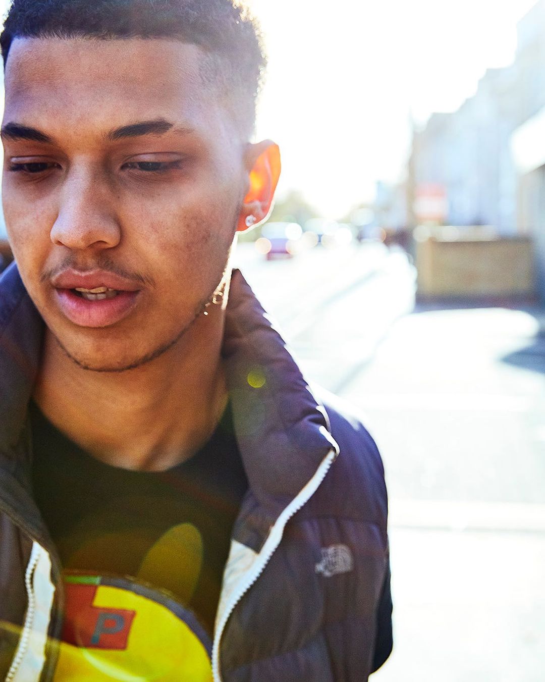 Close-up portrait of a young adult male outside in a street with cars and buildings in the background, bathed in natural sunlight.