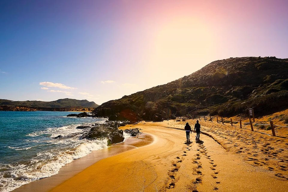 Two people walk along a sandy beach at sunset, with rolling hills, blue sky, and turquoise ocean waves visible in the background.