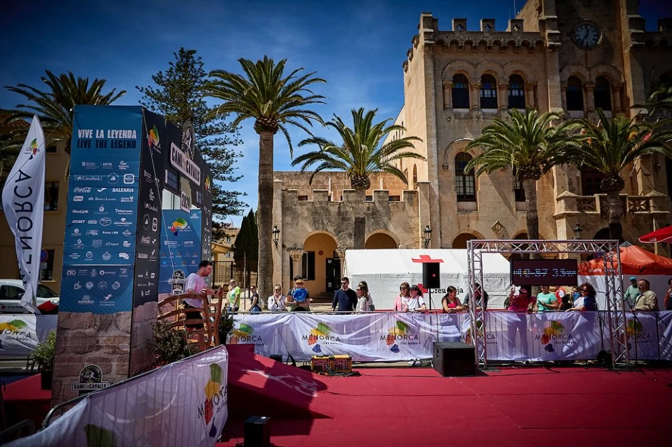 Outdoor event on a red carpet with a small crowd watching a man on a platform in front of a historic building and palm trees.