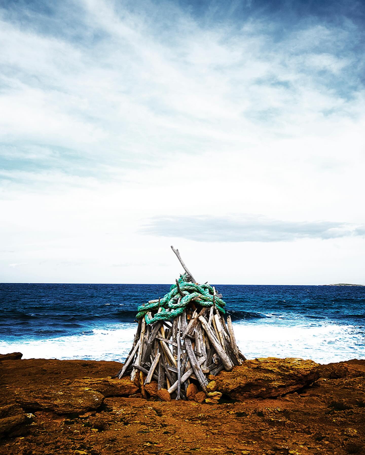 Driftwood shelter on rocky beach, ocean waves and horizon. Stack of wood with rope sits near blue water and cloudy skies.