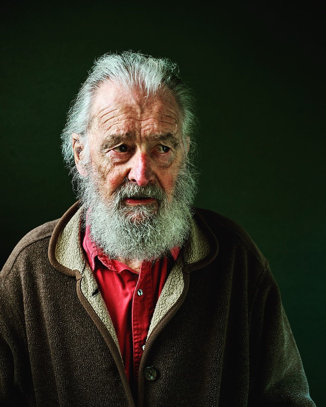 Portrait of an elderly Caucasian man with a long white beard, wearing a brown jacket over a red shirt, against a dark green background.