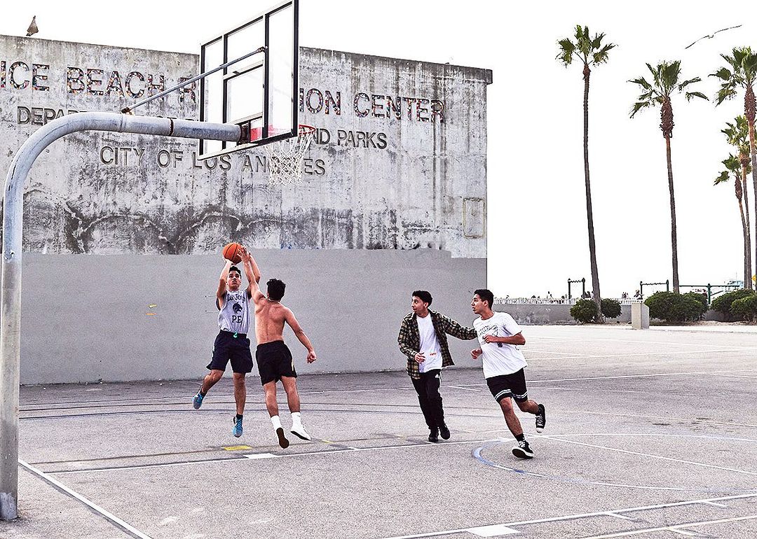 Four young adult men play basketball on an outdoor court on a sunny day near palm trees and a concrete building with text.