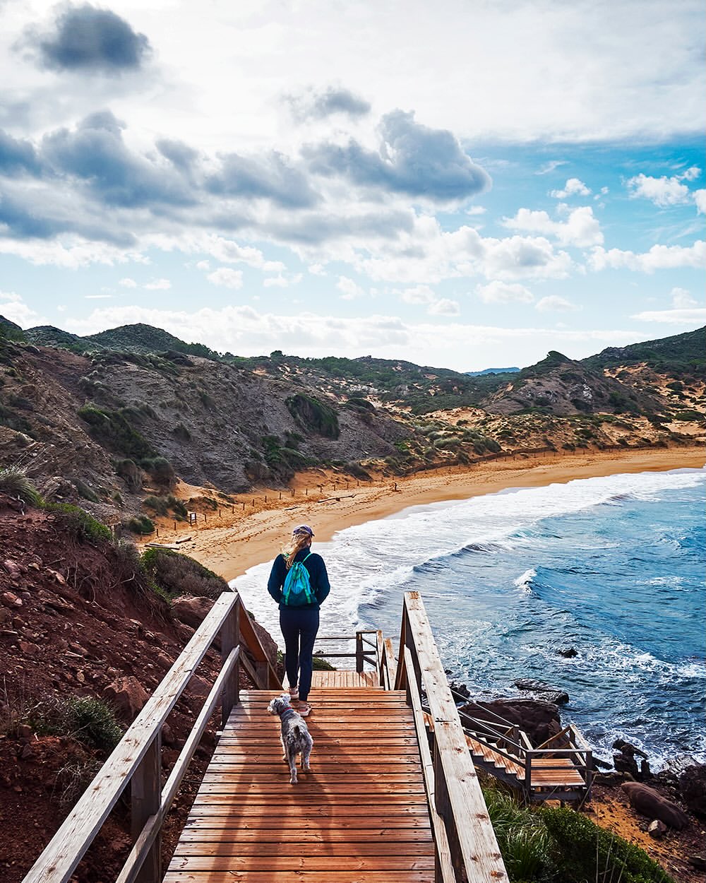 Woman with dog walking down wooden steps toward a beach with rolling waves, hills, and a cloudy sky.