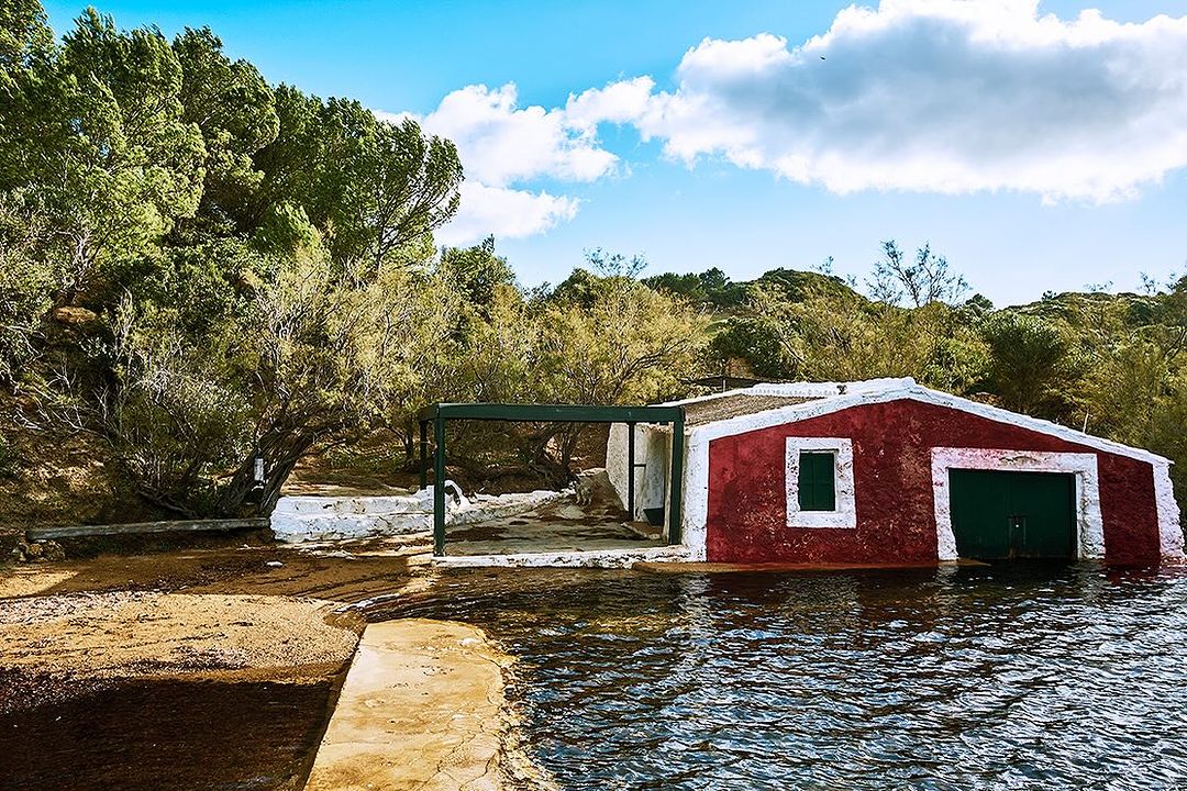 A red boathouse sits partially submerged in water near a sandy bank with lush green trees and a cloudy blue sky in the background.